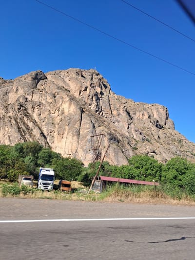 The image depicts a roadside scene in Malishka, Armenia, with a prominent rocky mountain dominating the background under a clear blue sky. In the foreground, a paved road with a white line runs horizontally. Beside the road, there is a collection of vehicles and structures. A white Iveco truck is parked centrally, to its left is a white pickup truck, and to its right is a rusty, possibly abandoned, van. Behind the vehicles, a line of green trees and vegetation extends across the landscape. Further back, a long, low building with a red roof and dark siding is visible, partially obscured by tall grass. A rusted utility pole with electrical wires is situated in front of the mountain. The mountain itself is steep and rugged, with visible rock formations and sparse vegetation. A small cross is visible on the summit of the mountain.