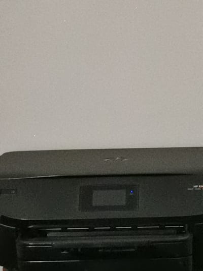 This is a close-up, low-angle, indoor, eye-level shot of the top portion of a black HP Envy printer. The printer has a flat top surface with a slight texture, and a dark, reflective strip running across its width. In the center of the printer's top surface, there is a small, rectangular display screen with a blue light illuminated in its center. To the right of the display, the text "HP EN" is partially visible. The background is a plain, light gray wall. There are no people, actions, or interactions depicted. The image appears to be taken in a home or office environment, and there is no information available about its specific location in Bourg-lès-Valence, France.