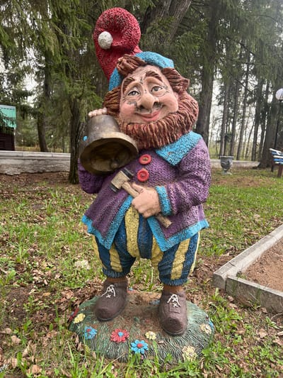 An outdoor, eye-level shot captures a colorful garden gnome statue set against a natural backdrop. The statue, depicting a whimsical dwarf with a reddish-brown beard, wears a red pointed hat with a white pompom, a purple jacket with blue trim and red buttons, and yellow and blue striped pants. Its face shows a slightly mischievous or inquisitive expression, with prominent ears and a large nose. The gnome holds a dark-colored bell in one hand and a small hammer in the other.
The statue stands on a green, grass-like base adorned with blue, red, and yellow painted flowers. The ground around it is covered with grass, patches of clover, and some scattered dry leaves. In the background, numerous coniferous trees, possibly pines or spruces, rise towards the sky, creating a dense, natural screen. A concrete border or planter is visible to the right of the statue, and a small, dark-colored structure, possibly a shed or building, can be seen further back among the trees. A single hanging light bulb is visible in the upper right background in one of the hint crops.
The scene appears to be set during daytime under an overcast sky or diffused light, as there are no harsh shadows. No specific activity is depicted beyond the static presence of the statue. The location is Sanatoriya Yumatovo Imeni 15-Letiya Bassr, Russia. There is no visible text in the image.