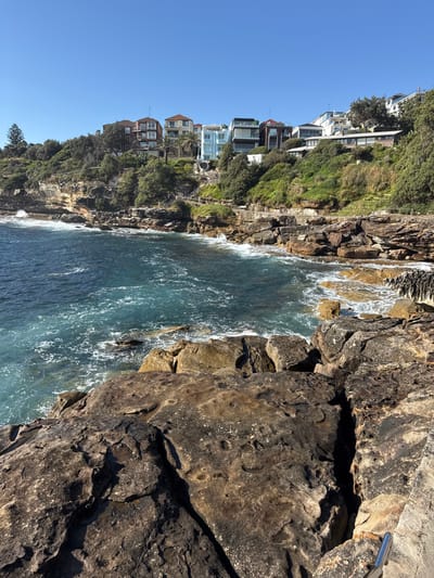 Rocky coastline meets the ocean under a clear blue sky. Buildings perch atop a green, rocky cliff overlooking the water. Waves crash against the rocks, creating white foam. The dark, textured rocks form a prominent foreground. A silver railing is visible in the top right corner.