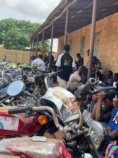 The image depicts an outdoor scene in Kassa, Nigeria, characterized by a gathering of people and numerous motorcycles parked under a covered structure. The structure appears to be a rudimentary building with a corrugated metal roof supported by wooden poles, adjacent to a brick wall. A large tree with dense green foliage is visible in the background.

In the foreground, several motorcycles are prominently displayed, some with visible political campaign materials. One motorcycle has a sticker featuring a man's portrait and text that reads "Late Mr. GWOM JERRY JOHY". Another motorcycle displays a sticker with the image of a man in traditional attire and the text "APC".

A group of individuals, predominantly men, are seen interacting and observing. Some are standing, while others are seated or leaning against the motorcycles. Their attire varies, with some wearing casual clothing and others in more formal or traditional garments. The mood appears to be somber or contemplative.

The environment suggests a local community setting, possibly a gathering for a political event or a community meeting. The presence of so many motorcycles indicates that transportation is a key element of this gathering, likely involving people from the surrounding area. The overall scene conveys a sense of community and political engagement within Kassa, Nigeria.