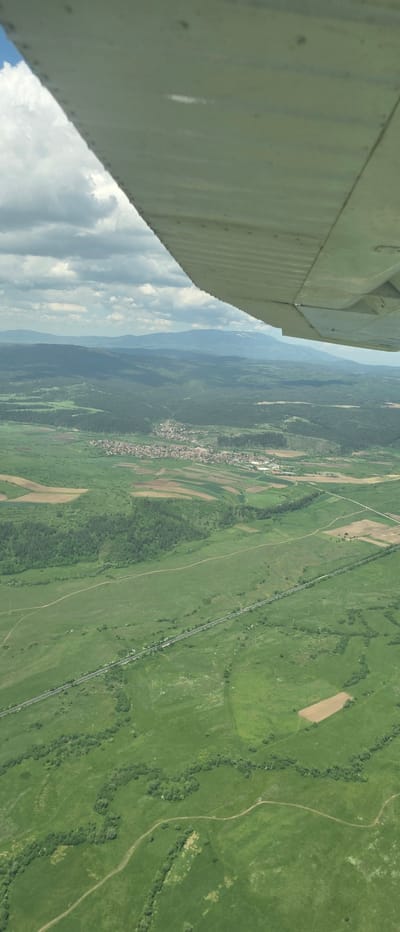 The image shows an aerial view of a landscape, likely taken from a plane or high vantage point. In the foreground, a portion of the aircraft's wing is visible, partially obscuring the view. The landscape below is composed of green fields, forests, and agricultural plots. A small village or settlement can be seen clustered along a linear feature, possibly a road or river. A road or railway stretches across the landscape. The location is the city of Dragushinovo, Bulgaria.