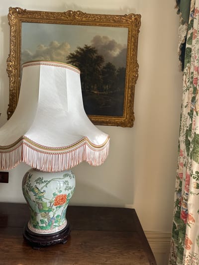 This image displays an interior scene featuring a collection of decorative objects. The primary subjects are a large, ornate table lamp with a white lampshade adorned with a tasseled trim and a Chinese porcelain vase. The vase, decorated with a detailed floral and bird motif, rests on a dark wooden base. To the left of the lamp, a framed landscape painting hangs on a cream-colored wall. The painting depicts a dramatic sky with dark clouds over a wooded area. Above the wall, a floral patterned valance with green, pink, and white flowers is visible, suggesting a window treatment. To the right of the vase, a portion of a dark wooden surface, possibly a piece of furniture like a desk or cabinet, is visible. The lighting suggests an indoor setting, possibly during the daytime with ambient light. There are no people or text visible in the image. The overall impression is one of a traditional, well-decorated room.