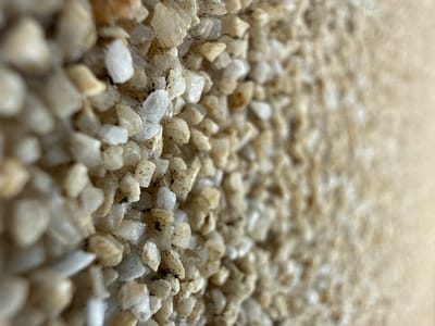The image displays a macro-level, close-up perspective of a textured surface composed of numerous small, irregular, and angular granular particles. These particles are predominantly light-toned, ranging from off-white and cream to light beige and translucent grey, with occasional darker flecks or adhered debris. The particles are densely packed and appear to be affixed to an underlying substrate, forming an uneven, rough surface.
The depth of field is shallow, with the sharpest focus on the particles in the left and central portions of the frame. Towards the right side, the granular texture rapidly blurs into an indistinct, lighter beige mass, suggesting a vertical or near-vertical plane receding from the viewer, or a surface where the focus gradually fades. No identifiable larger structures, objects, individuals, or specific environmental features are present within the frame that would allow for geographic identification. The image does not contain any visual or embedded metadata to determine its location in the city of Valky, Ukraine, or any other specific geographical coordinates. No actions or interactions are depicted.