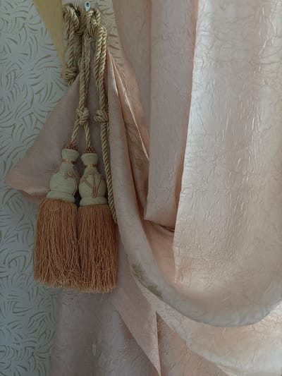 This image depicts an interior scene in the city of Velikiye Luki, Russia. A light pink curtain, characterized by a visible crushed or crinkled texture and subtle pale gold, possibly embroidered, leafy patterns, is draped and drawn to the left. It is held open by a decorative tie-back made of braided light gold rope. The rope forms two knots and is secured to a metallic, silver-toned wall hook located at the upper left. Hanging from the rope are two identical tassels, each featuring a contoured, gourd-shaped head wrapped in light beige cord with distinct peach-toned raised piping. Below each head, a voluminous fringe of fine, medium peach-brown strands extends downwards. To the left of the curtain and tie-back, a light-colored wall is visible, adorned with wallpaper that displays a repeating pattern of stylized, organic shapes in a subtle gold hue. The scene is illuminated by soft, diffused light, highlighting the textures of the fabric and wall covering. No individuals are present, and no specific actions are underway.