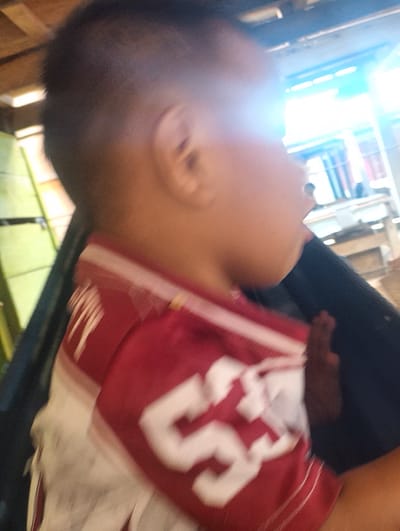 The image shows a close-up, blurred side profile of a young person, likely a child, with dark hair and light skin. They are wearing a red and white striped shirt with a large white number "53" prominently displayed on the front. The background is out of focus but appears to be an indoor or semi-outdoor setting with wooden structures and bright, possibly artificial, lighting. The overall impression is of a candid, motion-blurred photograph. Without further information, it is not possible to definitively place this image in Pompanua, Indonesia.