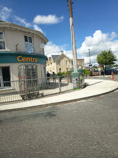 The image captures a street scene in Millstreet, Ireland, on a sunny day with scattered clouds. In the foreground, a blurred asphalt road suggests the perspective of a moving vehicle. A sidewalk curves along the right side, bordering a street and a grassy area.
On the left side of the image, a Centra convenience store is visible, with its signature orange "Centra" logo prominently displayed above a teal-colored facade. In front of the store, metal shopping trolleys are stacked. Two individuals, a man and a woman, are walking past the store on the sidewalk.
Further down the street, a tall utility pole stands prominently, with various wires and equipment attached. Behind the Centra store, two multi-story buildings with light-colored facades and cream-colored trim are visible. One building has a distinctive pointed roofline. A mature green tree stands to the right of these buildings, casting a shadow on the sidewalk.
Attached to a fence near the Centra store is a flyer advertising the "Millstreet Athletic Club Willie Neenan Memorial 5-Mile Road Race" scheduled for "Fri 26th June." The flyer details race times and registration information.
In the background, several parked cars can be seen, along with streetlights and traffic signals. The overall environment suggests a typical commercial and residential area in a small Irish town.