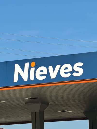 The image displays an upward-angled exterior view of a business sign under a clear blue sky. The prominent subject is a large, horizontal sign with a blue background, featuring the word "Nieves" in bold, white, rounded sans-serif letters. The dot above the 'i' in "Nieves" is distinctively orange.

Below the blue sign and an orange horizontal strip, the corrugated underside of a building canopy is visible, supported by two grey pillars. This structure suggests the location is a service station, such as a gas station, providing cover for vehicles or patrons.

Above the sign, the sky is a bright and clear blue, indicating a sunny daytime scene with no clouds. Three thin, dark power lines are stretched across the upper part of the sky, running horizontally.

The scene is static, showcasing the signage and parts of the building against the bright sky. The location is Sant Esteve Sesrovires, Spain.