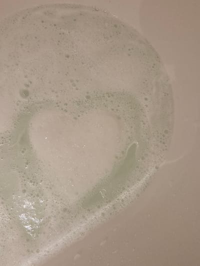 A top-down photographic view displays a quantity of light-colored, possibly pale green, foamy bubbles floating on a liquid surface within a light-beige or off-white receptacle, presumably a bathtub or basin. The foam primarily occupies the upper and left areas of the frame, forming an approximate heart-shaped void or area of reduced foam density in the central region. The surface beneath the foam appears smooth and uniform. No persons or objects other than the foam and liquid are present. The location of this scene is the city of Eeklo, Belgium.