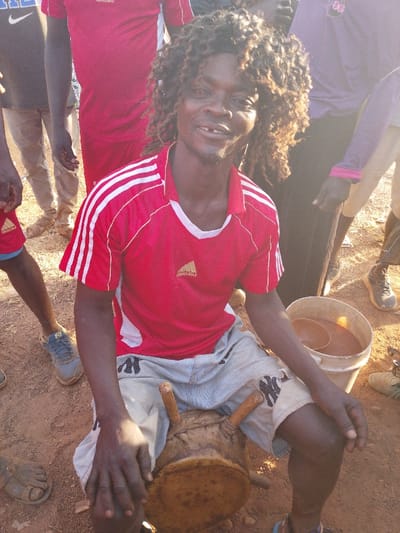 An outdoor scene in Vodni, Nigeria, depicts an adult male centered, seated amidst a group of partially visible individuals on dry, reddish-brown soil. The central male, with dark skin and dark, curly hair, smiles directly at the viewer, revealing a gap between his upper front teeth. He wears a red short-sleeved polo shirt with white stripes on the shoulders and an "BRISGDAS" (resembling Adidas) logo, paired with light-colored shorts patterned with dark lettering. He is seated on a small, crude, skin-covered drum with wooden protrusions, positioned between his legs, with his hands resting near it. To his right, a white plastic bucket contains a reddish-brown liquid with a smaller, lighter-colored bowl submerged. Surrounding the central figure are the legs and lower torsos of multiple other individuals, dressed in various casual attire including different colored shirts (red, purple, dark) and pants/shorts, and wearing diverse footwear such as sandals and sneakers.