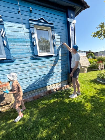 A full, eye-level outdoor shot depicts a man and a child painting a weathered blue wooden house on a sunny day in the village of Dunilovo, Russia. The house has decorative white trim around the windows. The man, wearing a grey t-shirt, grey shorts, a blue baseball cap, and white sneakers, stands to the right and paints near a window. He holds a paint scraper in his right hand, reaching up to the wall. A blue house number "22" is visible on a post to the right of the man. To the left of the man, a young child wearing a light brown onesie, a white patterned sun hat, and blue gloves, walks away from the camera. The child is holding a white object in their gloved hands. The house is set against a backdrop of a lush green lawn, a flower garden with purple and pink blooms, and a wooden well. The sky is clear and blue. The shadows of the house and people are cast across the lawn.