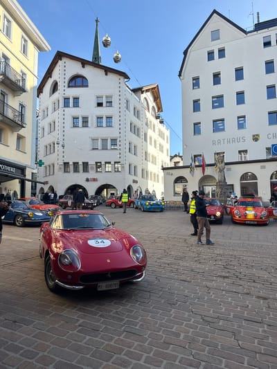 The image depicts a vibrant outdoor scene in Sankt Moritz, Switzerland, specifically a cobblestone town square surrounded by multi-story European-style buildings under a clear blue sky.

In the immediate foreground, a shiny red vintage sports car, identified as a Ferrari 275 GTB, is prominently displayed. It features a circular white race number "34" on its hood and has an Italian license plate "MI 6U0765." The cobblestone pavement extends from the foreground into the midground.

In the midground, several other vintage sports cars are parked or idling. To the left of the red Ferrari is a dark blue Porsche 911 with a yellow headlight cover and race number "38." Further back, a red and yellow striped car (likely an Alpine A110) with race number "39" has its driver's door open. Another red vintage car with race number "40" is positioned behind it. Additional vintage vehicles, including a light blue one and a red car with race number "36," are visible scattered throughout the square.

Numerous people are gathered around the cars, appearing to be spectators or participants in a car event. One individual in a yellow high-visibility vest stands near the red and yellow striped car, looking right. Another man in a dark jacket and brown pants stands nearby, looking at a mobile phone. People are generally observing the vehicles or engaging in conversation.

The background is dominated by distinct white buildings. On the right, a large white building is clearly labeled "RATHAUS" (City Hall) in grey letters, adorned with two coats of arms and flying Swiss and local flags near an arched entrance featuring a statue. Adjacent to the Rathaus, signs for "CHESA CUMUNELA" and "TAKE AWAY" are visible. To the center-left, another multi-story white building with distinctive stone detailing on its corners displays multiple "UBS" bank logos on its lower facade. Between these two main buildings, a tall, slender green spire, likely a church tower, rises towards the sky. On the far left, a light yellow building with dark wrought-iron balconies is partially visible, with "APOTHEKE" (pharmacy) visible on its ground floor. Decorative, cylindrical hanging lights are suspended across the square. The scene is illuminated by bright daylight, casting visible shadows.