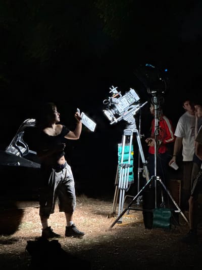 A person lies on a metal support, holding a clipboard, seemingly on a film set at night. Above them, a professional camera is mounted on a tripod. Further up, two other people are visible in the background. The scene is mostly dark, lit by a spotlight that casts a shadow on the ground. The surroundings suggest an outdoor, possibly wooded, location.