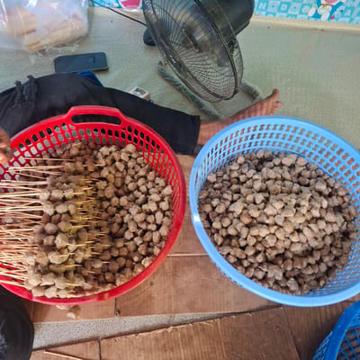 Two plastic baskets filled with small, brown, circular food items, likely meatballs or similar snacks, are positioned on a flat surface composed of cardboard and a textured floor covering. A red basket is on the left, containing skewers of these food items along with loose ones. A blue basket, on the right, is filled only with loose food items. A person's legs, clad in dark trousers, are visible behind the baskets, along with a black fan and a plastic bag containing food items in the background. A dark-colored mobile phone lies on the floor in the upper left portion of the image. The scene appears to be outdoors or in a semi-covered area, given the lighting and the type of floor. The location is identified as Reudeuep, Indonesia.