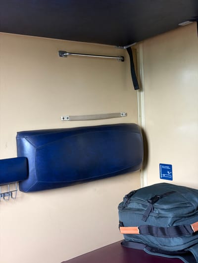 The image depicts the interior of a compartment on a train, likely in Chop, Ukraine, given the context. The compartment walls are a light beige color. A dark blue upholstered headrest or cushion is mounted horizontally on the wall. Above the headrest, a thin metal bar is attached to the wall, possibly for hanging clothes. A black fabric strap hangs down from the ceiling near the right wall. On the right wall, a small blue sign with white text and graphics is affixed, indicating some sort of instruction or information. In the foreground, a dark green backpack with black straps and tan leather accents rests on a dark red surface, which appears to be part of the train's seating or bedding. The lighting is somewhat dim, suggesting an indoor, possibly nighttime or overcast, environment.