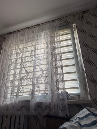 The image shows a window with white lace curtains and horizontal blinds. The curtains have an ornate floral pattern. A small white jar with a red label is visible on the windowsill. To the right of the window, wallpaper with a wavy pattern is present. The lower part of the image shows a radiator and a corner of a bed with striped bedding. The scene appears to be indoors, likely in a residential setting.
