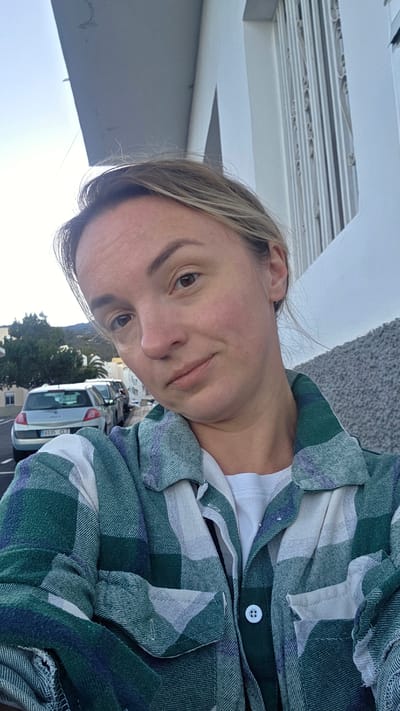This is a selfie taken by a woman outdoors in Guía de Isora, Spain. She is wearing a green and white plaid shirt over a white t-shirt. Her hair is blonde and pulled back. The background shows a street with parked cars, buildings, and trees, suggesting an urban or suburban setting. The sky appears bright, indicating it is daytime. The woman has a neutral expression. A white railing is visible in the foreground.