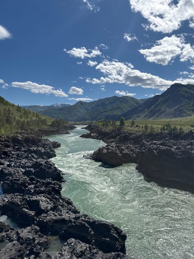 The image depicts a landscape near Edigan, Russia. A wide, rapidly flowing river occupies the central and right portions of the frame, its surface shimmering in the sunlight. Jagged, dark rocks form the foreground and the right edge of the riverbank. Mountains covered with sparse vegetation are visible in the background, under a blue sky dotted with white clouds. The image is rotated, so the sky is on the left.