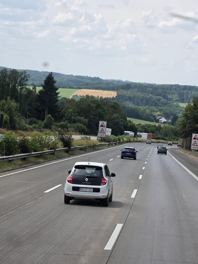 The image depicts a section of a German Autobahn, specifically near Oberhaid, Germany, on a cloudy day. The viewpoint is from a moving vehicle, looking forward and slightly to the right. Several cars are visible on the multi-lane highway. In the foreground, a white Renault Twingo is in the right lane, displaying the license plate NR-ZF 814. Further ahead, other vehicles, including a dark blue Audi, a silver hatchback, and a dark-colored sedan, are traveling in the same direction. The landscape on either side of the Autobahn consists of rolling hills covered in dense green forests, with fields of crops in the distance. Some agricultural buildings and houses are scattered throughout the rural scenery. Road signs indicating curves and distance are present along the roadside.