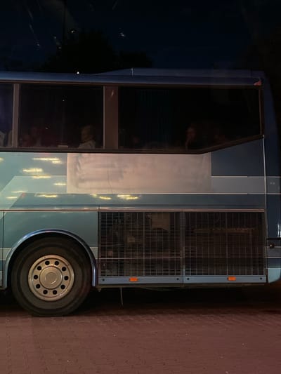 The image shows the side of a blue bus parked on a cobblestone street in Terespol, Poland. The bus has tinted windows, and through them, several people can be seen inside. The lighting suggests it is nighttime or dusk. The bus has a large wheel visible in the foreground, and a ventilation grate along its side. The street surface is made of interlocking cobblestones, and there are faint reflections of lights on the bus's exterior.