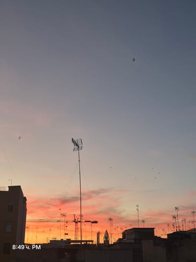 The image captures a vibrant sunset over the rooftops of Barletta, Italy, at 8:49 PM. The sky transitions from a pale blue at the top to a fiery orange and pink near the horizon, with wisps of clouds adding texture. Silhouetted against this dramatic backdrop are various urban elements. Numerous television antennas are visible, scattered across the rooftops, some standing tall on poles. A large construction crane is prominently featured in the mid-ground, its metallic structure stark against the colorful sky, suggesting ongoing development or renovation in the city. A building on the left, partially illuminated by the fading light, shows windows and architectural details. In the distance, the spire of a church pierces the sky, adding a touch of historical architecture to the modern urban landscape. Several birds are captured in flight against the expansive sky, adding a sense of movement and life to the scene. The overall impression is one of a city settling into the evening, bathed in the warm glow of a setting sun.