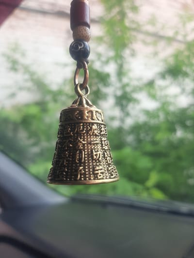 A close-up, eye-level, outdoor shot captures a small, ornate brass bell hanging from a string with beads. The bell is adorned with intricate carvings of what appear to be Chinese characters. Behind the bell, the background is softly blurred, showing out-of-focus green foliage and hints of a light-colored structure, suggesting it's near trees and possibly a building. In the foreground, a portion of a dark, reflective surface, likely a car's dashboard or window, is visible, indicating the bell may be a car accessory. The lighting is natural and somewhat diffused. The location is identified as Shelekhov, Russia.