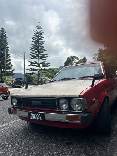 A vintage red Toyota Corolla is parked on an asphalt surface in a parking lot in Tanah Rata, Malaysia. The car's hood appears to be aged and discolored, contrasting with the otherwise bright red paint of the vehicle. The car has a license plate that reads "WW 3584." In the background, several tall evergreen trees with a distinctive conical shape are visible, along with other vehicles, including a black SUV and an orange compact car. The sky is overcast with dramatic, dark clouds, suggesting a potential for rain. The scene is framed by lush green foliage and hilly terrain in the distance, typical of the Cameron Highlands region. The overall atmosphere is one of a natural, slightly remote, and cloudy outdoor setting.