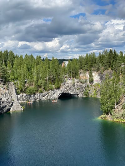 The image depicts the Marble Canyon in Ruskeala, Russia, a former marble quarry now filled with turquoise water. The canyon walls are steep and rocky, covered with lush green trees and vegetation. The water is dark blue and appears calm, with a few small boats with people in them scattered across its surface. In the distance, a cave entrance is visible in the canyon wall. The sky is overcast with dramatic clouds, creating a moody and atmospheric scene. There are people visible on the rocky ledges above the water, observing the canyon and the boats. The overall impression is one of natural beauty and a popular tourist destination.