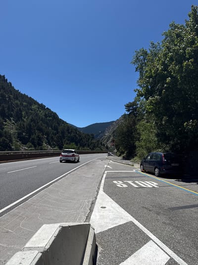 The image depicts a road winding through a mountainous landscape under a clear blue sky, suggesting a sunny day. The perspective is from a roadside, looking down the road. On the right side of the road, a concrete barrier separates the asphalt from a drop-off. White lines mark the lanes, and a "STOP" sign is painted on the road surface.
A dark-colored car is parked on the left side of the road, near the top of the frame, partially obscured by lush green trees that extend from the mountainside. Further down the road, two cars are visible: a white and red car in the right lane and a dark car behind it. The mountains are covered in dense evergreen trees, characteristic of a forest.
The presence of the cars and the road suggests a travel or transit scene. The clear sky and bright sunlight indicate daytime, likely late morning or early afternoon. The location cues, such as the mountainous terrain and vegetation, are consistent with Canillo, Andorra. No specific emotions are conveyed in the image, which focuses on the scenery and the road infrastructure. The visible text includes the word "STOP" painted on the road. The license plate of the parked car is partially visible, showing "CH 842 GH".