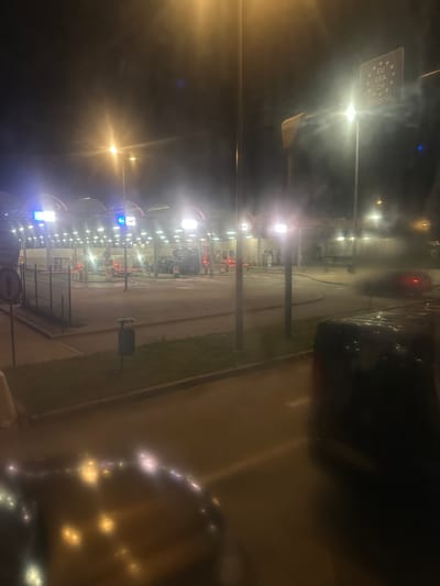 At night, a border crossing point is depicted in Hrebenne, Poland. The scene is illuminated by numerous bright lights, including streetlights and lights from the border crossing structures. Several vehicles are visible, including cars and a larger, dark-colored van in the foreground. Some cars are in motion, as indicated by blurred taillights. The border crossing area features covered lanes with traffic barriers and signs. A sign on a pole displays stars and text that reads "EU," "EEA," "CH," and "CITIZENS," suggesting it is an EU border crossing. A trash bin is situated on the side of the road. The overall environment appears to be an active, operational border checkpoint at night, with cars passing through.