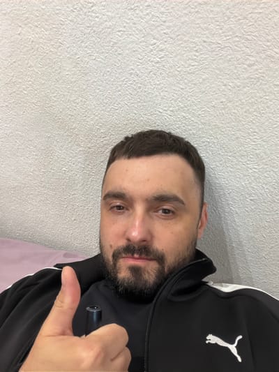 A man with short, dark hair and a beard is posing for a selfie indoors. He is wearing a black Puma jacket with white accents. He is holding a small black object in his hand and giving a thumbs-up. The background is a textured white wall. The location in the city of Kovel, Ukraine is not discernable from the image.