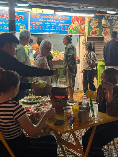 The image depicts an active outdoor night street food scene in Chiang Rai City Municipality, Thailand. In the foreground, a young woman wearing a black and white striped shirt is seated at a small, yellow square metal folding table. To her left, a man in a black long-sleeved shirt stands, serving a steaming liquid from a clay hotpot, resting on a glowing charcoal stove, into a green bowl which he holds. On the table, various dishes are present, including a large platter of greens and other food items in clear plastic containers, along with multiple plastic cups containing beverages, ice, and straws. A yellow bottle and a small metal ice bucket are also on the table. A smartphone stands upright on the table. To the right, another woman, dressed in a dark long-sleeved top, is seated, engrossed with a smartphone in her hands.
The background features a food stall with prominent blue and green illuminated signs displaying menu items in Thai and English, including "Hotpot Soup & Soup Spicy." Images of prepared dishes are visible on the signs. Beneath the signs, a counter displays an assortment of raw ingredients, likely for hotpot. Several individuals are standing in the mid-ground near the food stall: an older man with grey hair, an older woman with white hair and glasses looking towards the foreground, a man with a bun hairstyle, a woman in a patterned sweater, and another man with his back partially to the camera. The area is brightly lit by overhead artificial lights emanating from the food stall, and the ground surface exhibits a patterned tile design.