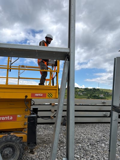 In Skrzydlna, Poland, a construction worker wearing a white hard hat and an orange safety vest is operating a yellow scissor lift. The worker is positioned at the top of the lift, which is elevated to a height level with a section of steel framing. The worker appears to be attaching or securing the steel frame, holding a tool that resembles a cordless drill or impact driver. In the background, a hilly landscape is visible with scattered houses and trees under a partly cloudy sky. A pile of stacked steel beams lies on the ground in the mid-ground. The ground is covered in gravel.