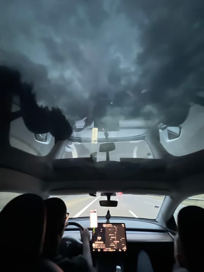 An interior, low-angle, wide shot from the back seat of a modern electric vehicle, possibly a Tesla, shows the occupants as dark silhouettes. The car's panoramic glass roof reveals a dark, cloudy sky, suggesting overcast or stormy weather. Ahead, the dashboard is visible with a large central touchscreen displaying a map. The driver, with glasses on, is seen from the side, gripping the steering wheel. The road ahead is illuminated and appears to be a highway. The context of Les Houches, France, is not discernible from the visual information presented in the image.