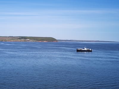 The scene depicts a body of water, presumably a river or lake, under a clear, slightly clouded sky. A ferry navigates the water, leaving a visible wake. In the background, a landmass with a distinctive reddish-brown cliff face topped with green vegetation is visible. The location is Икское Устье, Russia.