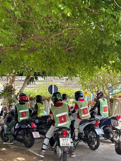 The image shows a group of people on motorbikes, likely in the city of Nong Phueng, Thailand. The individuals are wearing numbered vests (1, 2, 3, 4) and helmets. The motorbikes are parked next to each other, and license plates are visible on the vehicles. A tree is located to the left, providing some shade to the area. A building with a white-framed structure is visible in the background.