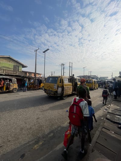 In Akowonjo, Nigeria, a sunny day with scattered clouds illuminates a busy street scene. Several yellow minibuses and auto-rickshaws, common forms of public transport, are parked and in motion. A group of people, including children, are visible on and around the vehicles, suggesting a moment of transit or waiting. The architecture consists of modest buildings, some appearing weathered, lining the road. Power lines crisscross the sky, a common feature of urban environments. The ground is paved and appears worn. The overall impression is of a dynamic, everyday street life in a Nigerian city.