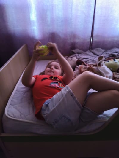 In the provided image, a young girl with blonde hair is lying on a mattress inside what appears to be a bed frame. She is wearing a red t-shirt with a colorful graphic and denim shorts. The girl is holding a small, yellow object, possibly a toy, in her hands. Her legs are bent and crossed.
The setting is indoors, with a purple, possibly fabric, backdrop behind the girl. To the right of the girl, on the mattress, are some blankets and a plush toy that has a yellow head and green ears. The lighting in the room is dim, with some natural light filtering through the purple material, casting shadows and highlights. The overall atmosphere appears relaxed and casual. There is no visible text in the image. The time of day and weather are not explicitly indicated, but the indoor setting and dim lighting suggest it could be evening or a cloudy day. The location context is Novy, Russia.