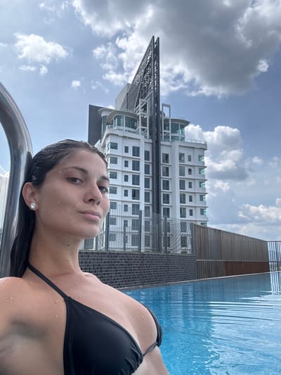 The image displays a woman relaxing in a swimming pool, with a modern cityscape as a backdrop. The photograph is taken from a low angle, rotated counter-clockwise, with the woman's head to the left and her body extending to the right.
The primary subject is a woman, partially submerged in the clear blue water of what appears to be a rooftop or infinity pool. Her head, with wet dark hair, is resting on a metallic pool edge or railing. Her face is turned towards the camera, showing a calm, slightly contemplative expression. Water droplets are visible on her skin, suggesting she has just emerged or is submerged. She wears a black bikini top and a small, round, sparkling stud earring in her visible ear.
In the background, a tall, contemporary high-rise building dominates the scene under a partly cloudy sky. The building is predominantly white or light grey, featuring numerous windows and distinct architectural elements, including a dark, lattice-like structure on its upper-left side and sections with curved glass at the top. Adjacent to the pool on the right, there is a wall or structure made of dark, possibly slatted or tiled material, complementing the modern aesthetic. The sky above is bright with scattered white and grey clouds.
The setting suggests an urban environment, likely a hotel or residential complex with an elevated pool, consistent with the provided context of Ampang Jaya Municipal Council, Malaysia. The time of day appears to be daytime, given the bright illumination. No visible text is present in the image.