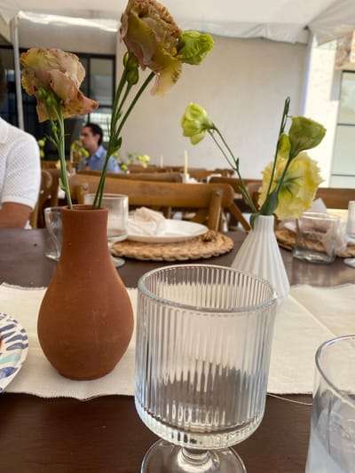 The image depicts a table setting, likely for a meal, located in Santa Cruz Tehuixpango, Mexico. The table is covered with a light-colored linen runner, and several place settings are visible. One place setting includes a blue and white patterned plate and a cylindrical ribbed glass. A small terracotta vase holds light pink and green flowers. Another vase, white and ribbed, holds yellow and green flowers. Behind the white vase are additional objects, including a wicker placemat, a white plate, and glassware. Further in the background, a person wearing a blue shirt is visible amidst the other table settings and decorations. The environment appears to be an outdoor or semi-outdoor space, possibly under a tent or canopy. The table itself is made of dark wood.