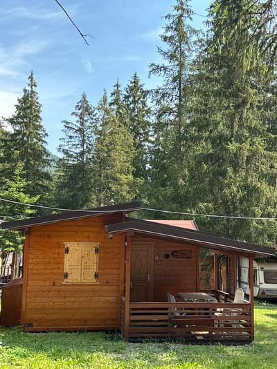 A wooden cabin with a covered porch is situated in a grassy area, surrounded by tall evergreen trees. A picnic table and chairs are visible on the porch, and a caravan is parked in the background. The sky is blue with some scattered clouds. The scene appears to be a campsite or a rural setting.