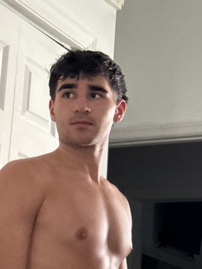 The image shows a young, shirtless man with dark, curly hair, looking to his right. He has dark eyebrows and eyes. He is standing in an indoor setting, with a white door to his left and a gray wall and a dark television screen in the background. The lighting appears to be artificial, creating shadows on his chest and shoulders. There is a white molding or trim visible at the top of the image. The overall scene suggests a casual or personal moment captured in what appears to be a residential environment within Bellmore, New York.