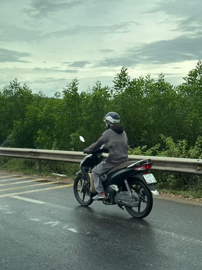 A person wearing a helmet, mask, grey hooded sweatshirt, and grey pants rides a black motorcycle on a wet asphalt road. The road has yellow and white striped markings. To the left of the road is a guardrail made of metal beams supported by concrete posts, beyond which is a grassy area. In the background, there is a dense line of green trees under a cloudy sky. The license plate on the motorcycle reads "74-F1 336.10". The scene appears to be in a rural or suburban area of Nam Hải Lăng Commune, Vietnam, given the presence of lush vegetation and the style of the motorcycle and its license plate.