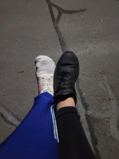 The image shows two pairs of shoes on a concrete surface with cracks. One pair is black athletic shoes worn with black pants. The other pair is light-colored athletic shoes with blue pants featuring white stripes. The setting appears to be outdoors, possibly a sidewalk or pavement, in Altdorf (UR), Switzerland, judging from the prompt.