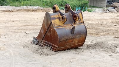 An excavator bucket is partially submerged in loose sand. The bucket is made of rusted metal with yellow paint markings. It has a wide, angled opening with several teeth or blades along the bottom edge. Two large metal pins connect the bucket to a pivot mechanism at the top, which is also yellow and appears worn. The bucket is resting on a patch of granular material, possibly gravel or coarse sand. The surrounding environment is a construction or undeveloped area, characterized by large expanses of light-colored sand with tire tracks visible in places. In the background, there are piles of dirt and debris, some scattered rocks, and a dense line of green vegetation. A concrete structure is partially visible in the far background. There are no people or animals present in the image. The scene appears to be outdoors during daylight. The location is identified as Beunyot, Indonesia.