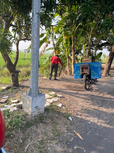 In Karangtanjung, Indonesia, a man wearing a red shirt and dark pants sweeps the ground with a broom near a metal pole. To his right is a blue box attached to a motorcycle. The box is decorated with images of shoes and has text in Indonesian, including "Cepat" and "Tarip Biasa." The surrounding environment is rural, with trees, vegetation, and a field of crops in the background. The ground is dirt and debris.