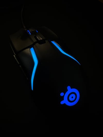 A black gaming mouse with blue LED lighting is positioned on a dark surface. The mouse has a sleek, ergonomic design with glowing blue accents along its sides and at the top near the scroll wheel. The SteelSeries logo, also illuminated in blue, is visible on the palm rest area of the mouse. The background is entirely black, indicating a dimly lit or dark environment. The image is taken from a top-down perspective, focusing on the mouse. There is no discernible location in the city of Mariannelund, Sweden mentioned or visible in the image.