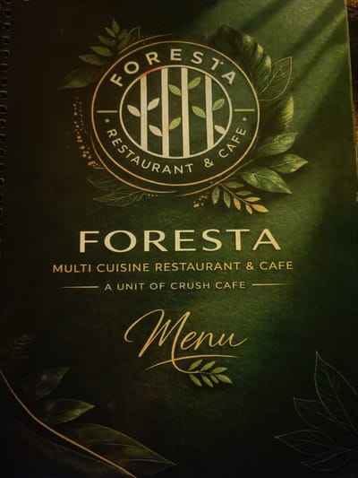 This is a close-up, eye-level shot of the cover of a restaurant menu. The menu is dark green with gold lettering and ornate graphics. The top of the cover features a circular logo that says "FORESTA RESTAURANT & CAFE" in gold lettering. Inside the circle are white vertical lines resembling bamboo stalks, with small green leaves. The rest of the cover is decorated with green leaves and gold accents. Below the logo, the words "FORESTA" are written in large gold letters, followed by "MULTI CUISINE RESTAURANT & CAFE" and "A UNIT OF CRUSH CAFE". At the bottom, the word "Menu" is written in a stylized gold script. There are no people or actions depicted. This image is likely from a restaurant in Kurnool, India, as indicated by the request.