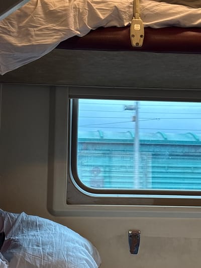 The image depicts the interior of a train compartment, likely a sleeping berth. A white sheet is draped over a dark maroon padded surface, which is likely the top bunk. A metal latch mechanism is visible on the maroon surface. Below this, a window with a rounded corner offers a view of the outside. The window is partially obscured by the movement blur of passing objects. Through the window, the sky appears blue, and there are horizontal lines suggesting structures or the landscape passing by. Below the window, a crumpled white sheet is visible, likely part of the lower bunk bedding. A small metal fixture, possibly a handle or latch, is attached to the wall below the window. The overall impression is of a utilitarian and somewhat confined space. The location in Nazar'evo, Russia, is not discernible from the visual information present in the image.