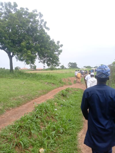A group of men, dressed in various traditional Islamic attire, are walking single file on a dirt path. The path is bordered by lush green grass. In the background, there are several houses with orange tiled roofs, and a large, leafy tree dominates the left side of the frame. The sky is overcast. The image appears to be taken in a rural or semi-rural area, consistent with some parts of Ife, Nigeria.